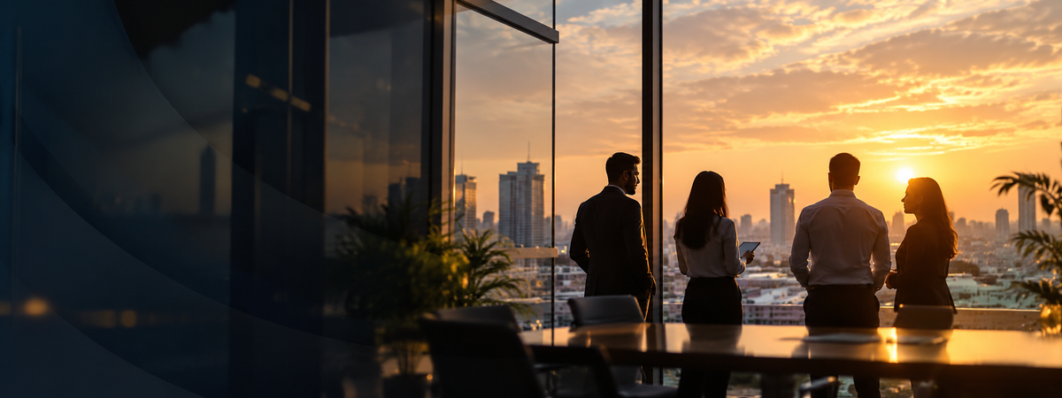 Silhouetted business professionals standing by a large window at sunset, overlooking a city skyline. The scene is warmly lit with golden hour light, and the office interior is visible in the foreground.