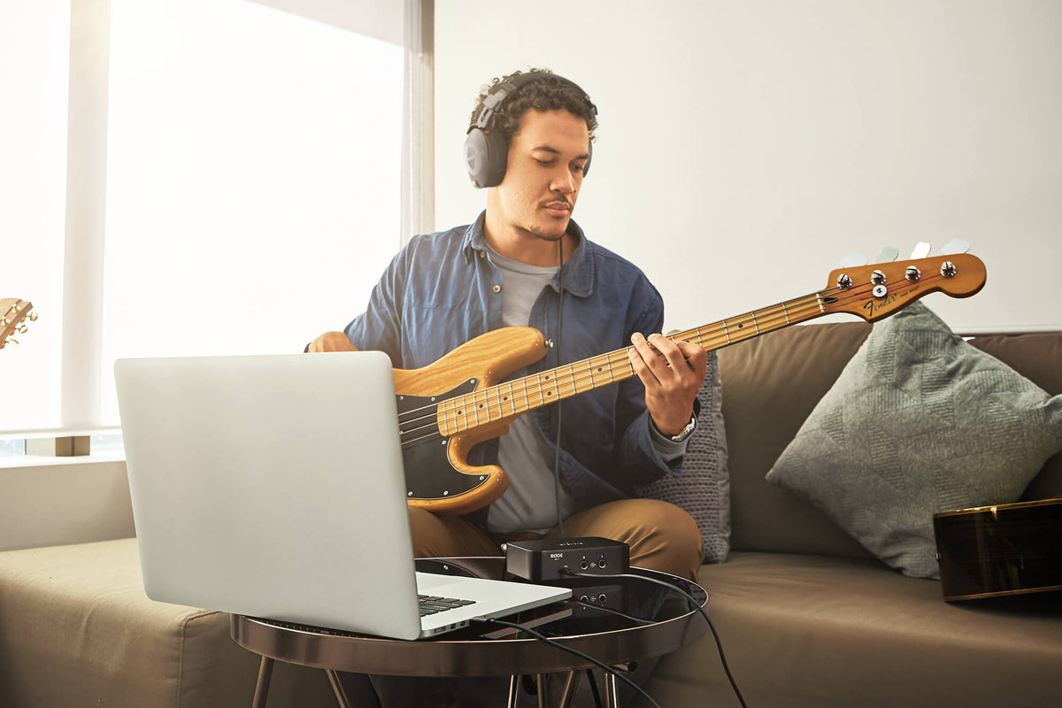 Musician with headphones playing bass guitar on a couch with a laptop and audio interface