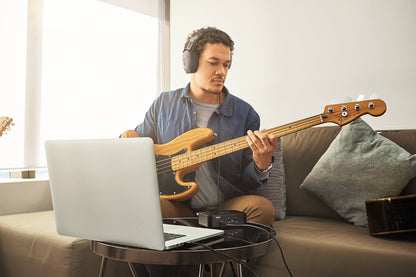 Musician with headphones playing bass guitar on a couch with a laptop and audio interface